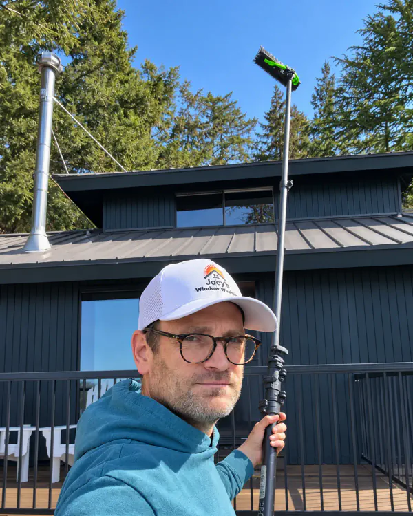 Joey in a cap and glasses, holding a water-fed cleaning pole in front of a house.
